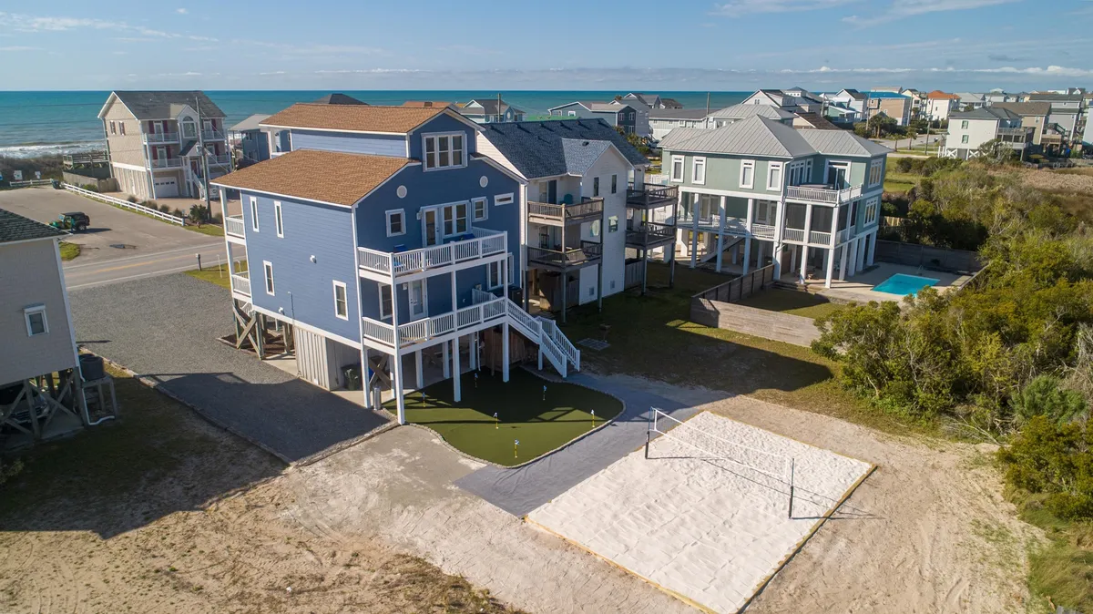 This exterior image highlights a beach volleyball court in front of a charming blue house, with a swimming pool visible and the ocean in the background. The scene is alive with the possibility of group activities and family gatherings, set against a backdrop of sunset hues and an open, inviting beachfront environment.