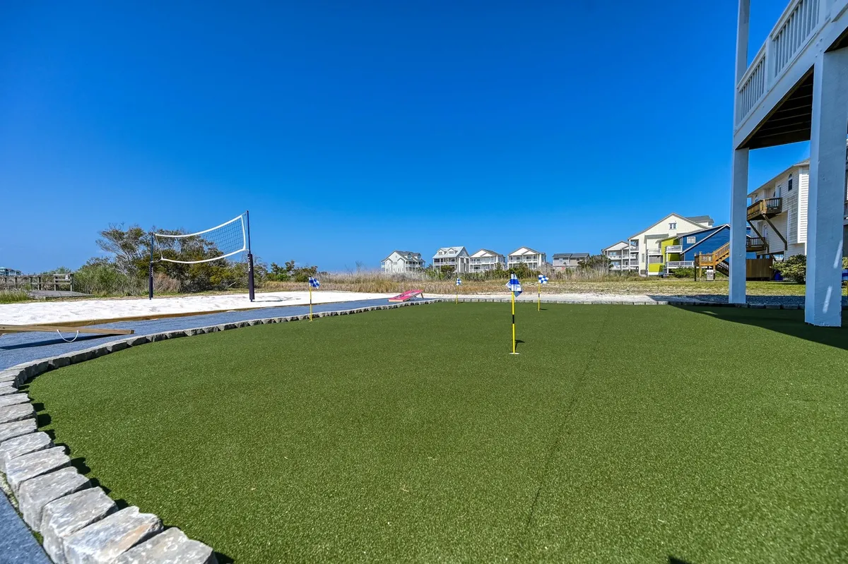 An inviting outdoor scene showing the putting green and volleyball net, with beach houses in the background and space for group gatherings. The scene is active and recreational, suggesting both daytime and sunset play.