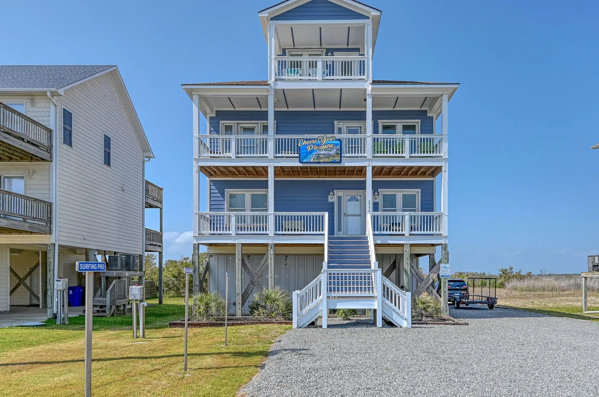 The image showcases a spacious three-story beach house with inviting front steps, multiple balconies perfect for relaxing, and visible signage, all set against the backdrop of North Topsail Beach. The home's architecture and large porches emphasize outdoor living, while the coastal setting hints at nearby ocean and intracoastal vistas.