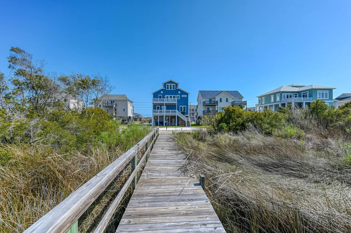 The blue house is connected to the outdoors by a wooden pathway winding through greenery, with the beach visible just beyond. The scene evokes the easy transition from home to beach adventures.
