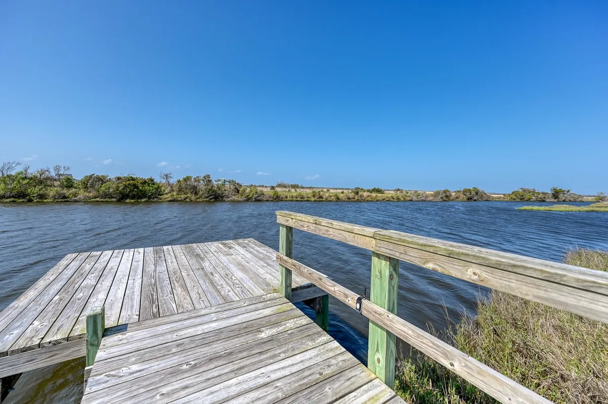 A peaceful wooden dock stretches over calm water, bordered by lush marsh greenery, inviting moments of relaxation or launch into kayaking and paddleboarding. The tranquil mood is heightened by soft sunset light reflecting on the water.