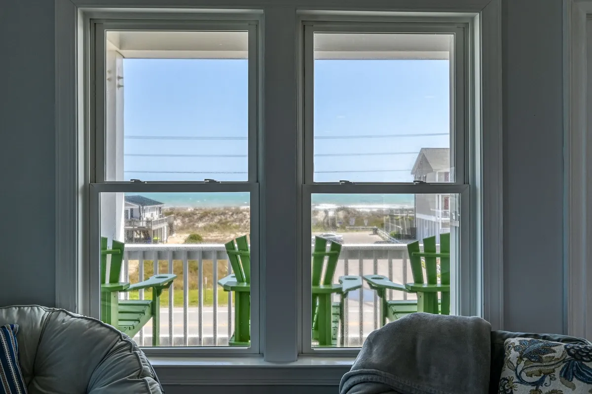 An ocean-view porch with green chairs overlooks the Atlantic, seamlessly blending the interior seating with the outdoors. The scene exudes tranquility, with an inviting atmosphere for relaxing, reading, or enjoying the sea breeze. The porch visually connects the living area to the spectacular water views.