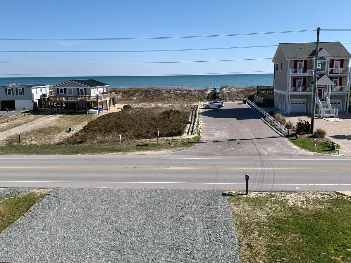 This exterior image shows two houses with an unobstructed view of the Atlantic Ocean. The foreground includes a roadway and parking area, providing a sense of proximity and easy access to the beach just across the street. The porch and surrounding area suggest relaxed family gatherings, while the ocean in the background hints at endless opportunities for beach outings and water activities.