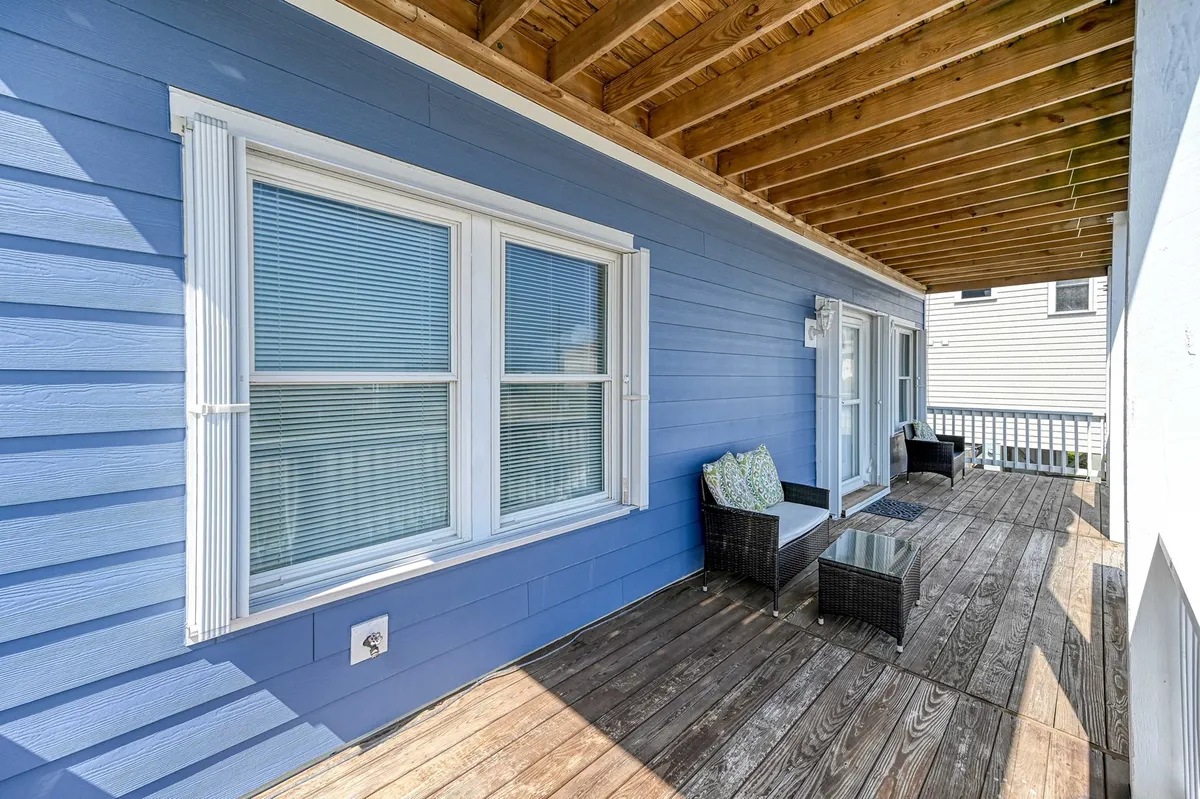 A tranquil outdoor deck featuring comfortable seating and decorative cushions, set against the home's blue siding. This covered porch area is perfect for relaxing with pets, enjoying a morning coffee, or unwinding together after a day at the beach. It provides shelter and a seamless indoor-outdoor connection.