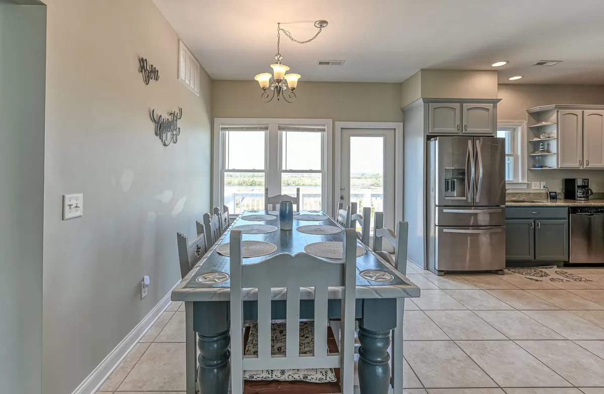 A long dining table sits adjacent to large windows, allowing natural light to flood the space. The table is surrounded by chairs, set for a family meal or gathering, and a chandelier above adds warmth and elegance. The kitchen and appliances are visible nearby, reinforcing the open-plan flow.